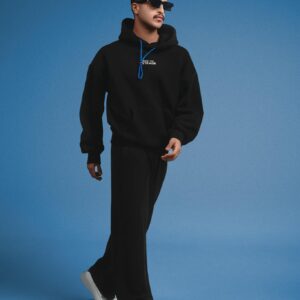 Fashionable man in black hoodie and pants walking against a blue backdrop.
