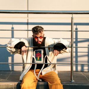 Fashionable man in yellow pants and colorful sunglasses sitting on a metal railing outdoors.