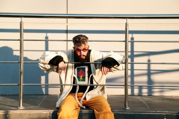 Fashionable man in yellow pants and colorful sunglasses sitting on a metal railing outdoors.